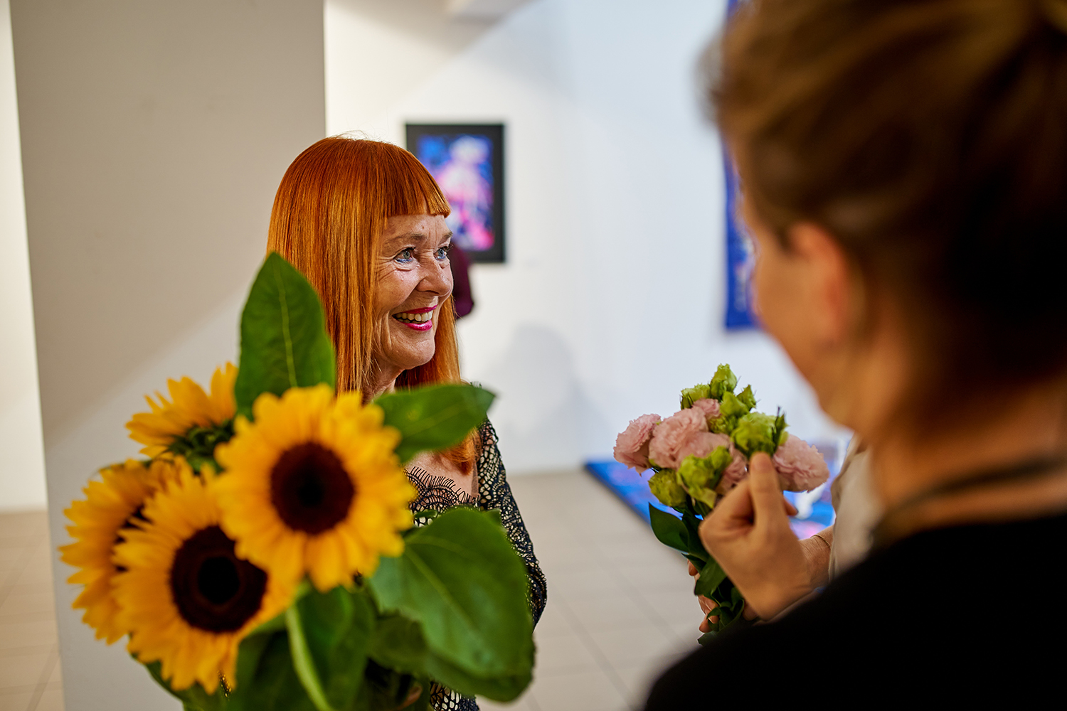Uśmiechnięta Julie Oakes, stoi na tle zaprezentowanych prac. Trzyma ogromny bukiet słoneczników i uważnie słucha osoby wręczającej jej kwiaty.