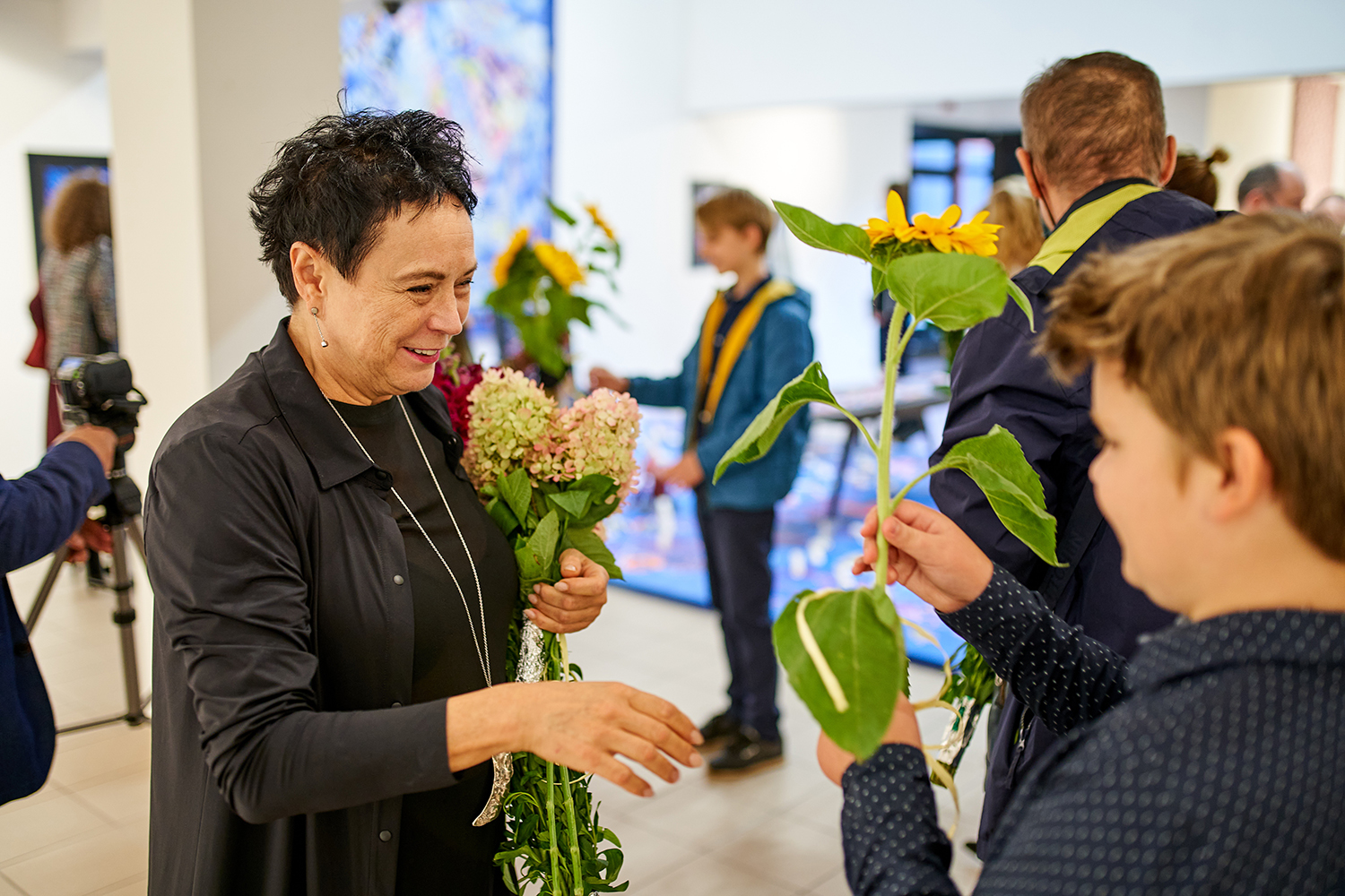 Na pierwszym planie prof. Jolanta Rudzka-Habisiak. Stoi w centrum kadru z naręczem kwiatów, na tle prac i zwiedzających. Rozpromieniona serdecznym  uśmiechem, przyjmuje  duży kwiat słonecznika od młodego chłopca, który odwzajemnia życzliwe gesty.