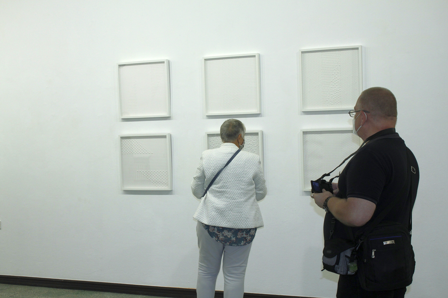 Kilka zwiedzających wystawę osób, na tle jasnego wnętrza galerii.  Widać tu sześć kwadratowych, białych prac rozwieszonych w dwu rzędach. Światło wydobywa strukturę prac i czyni ją dobrze widoczną nawet z dużej odległości.