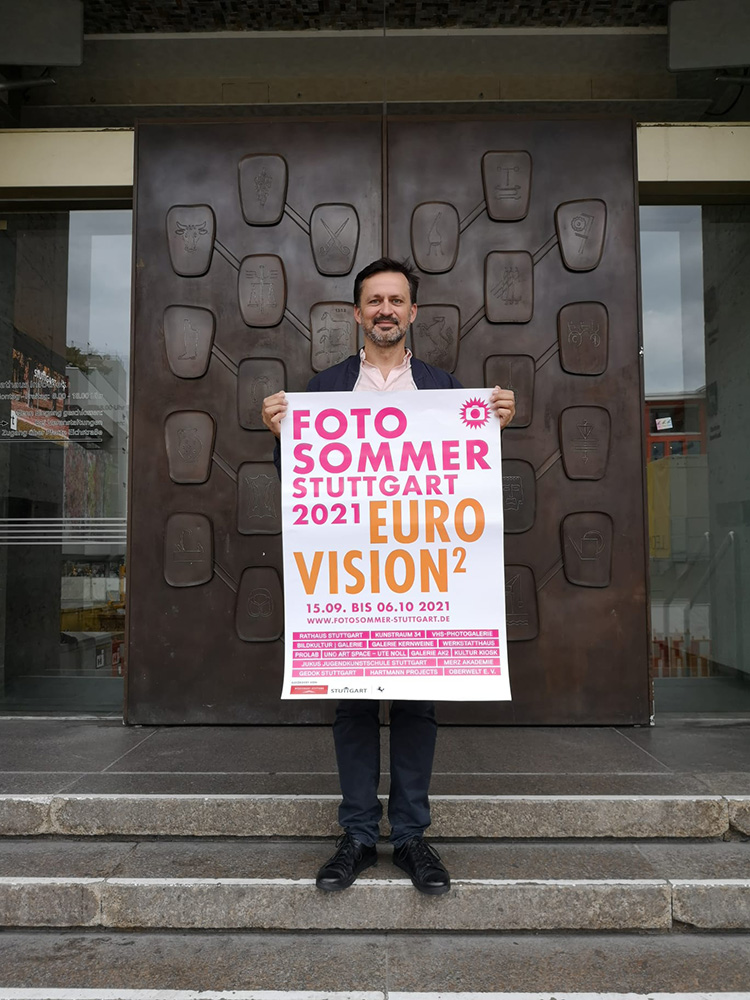 Zdjęcie wcześniej opisanego mężczyzny. Tutaj trzyma przed sobą w rękach plakat FOTOSOMMER STUTTGART 2021 EUROVISION 2, który został opisany na zdjęciu pierwszym. Postać stoi na tle ciemnobrązowej płaskorzeźby