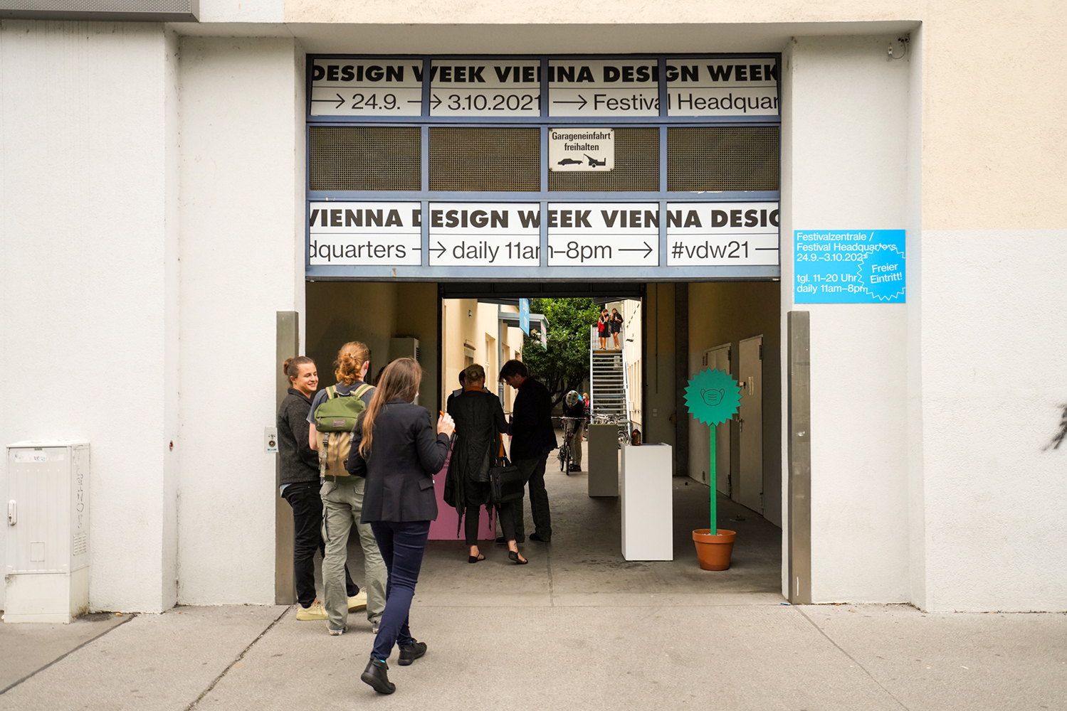 Szerokie wejście, nad którym widnieje napis: DESIGN WEEK VIENNA DESIGN WEEK 24.9-3.10.2021 FESTIVAL. Wokół budynku i widzianego w głębi patio stoi  wiele młodych osób zajętych rozmową