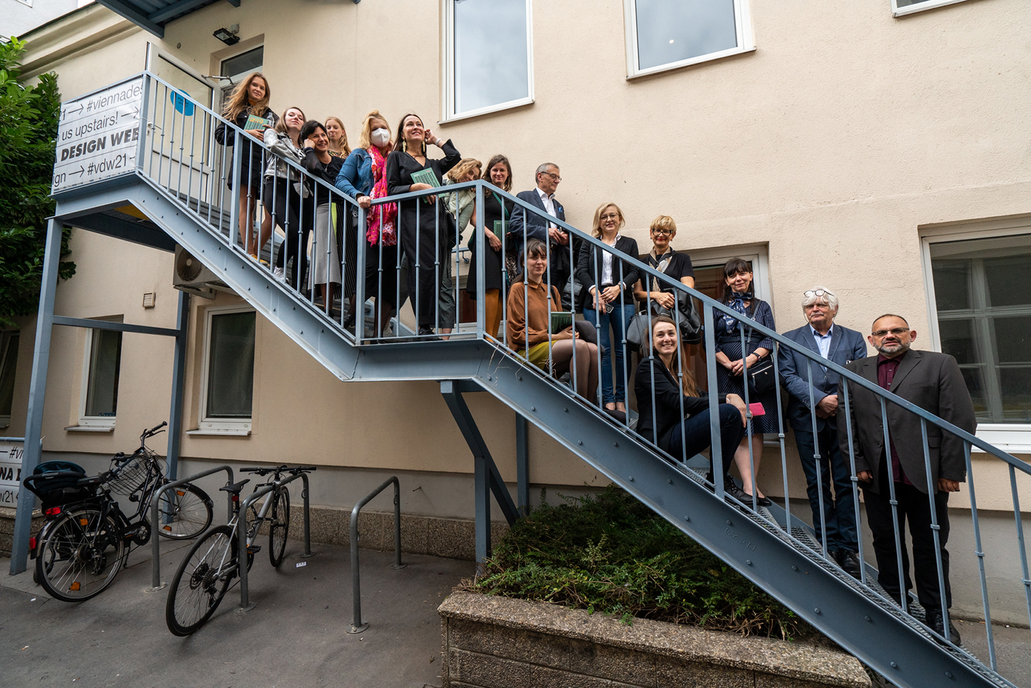 Na zewnętrznych metalowych schodach w kolorze granatu, wiodących na piętro beżowego budynku pozuje do pamiątkowego zdjęcia około dwadzieścioro osób, prawdopodobnie uczestników wystawy. Wszyscy są uśmiechnięci