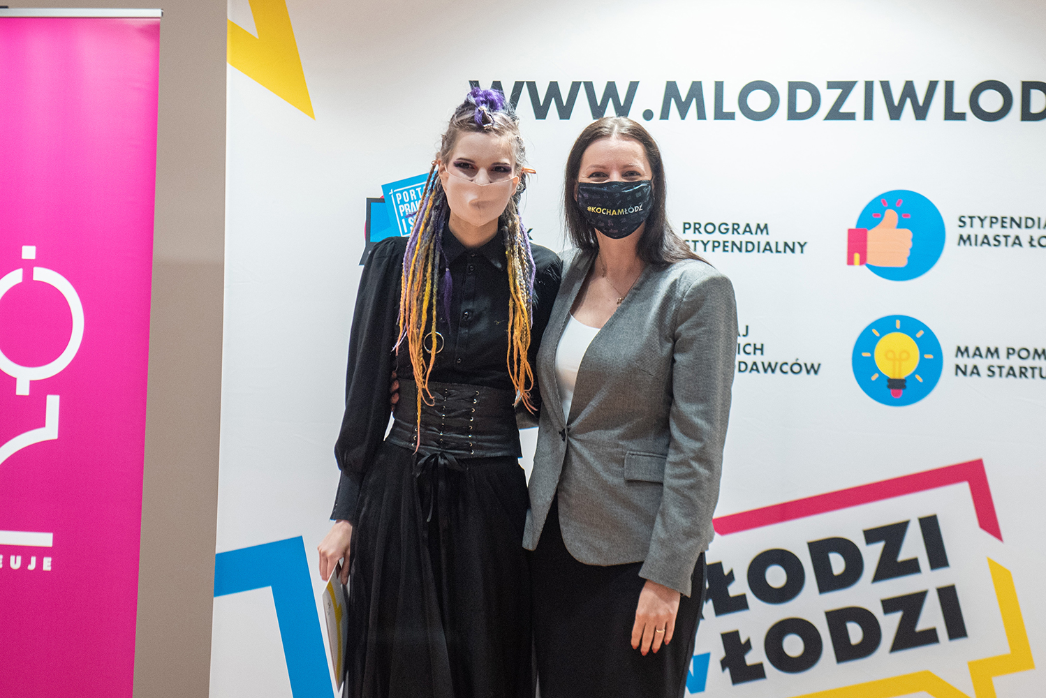 Na tym samym tle jak poprzednio, dwie kobiety. Miejsce młodszej zajęła inna, w podobnym wieku, także ubrana na czarno, z ciemno blond długimi włosami, uformowanymi w  pomarańczowo fioletowe dredy. Także i ona trzyma w prawej dłoni sztywną białą teczkę z nagrodą. Obok stoi ta sama kobieta  w szarym żakiecie.