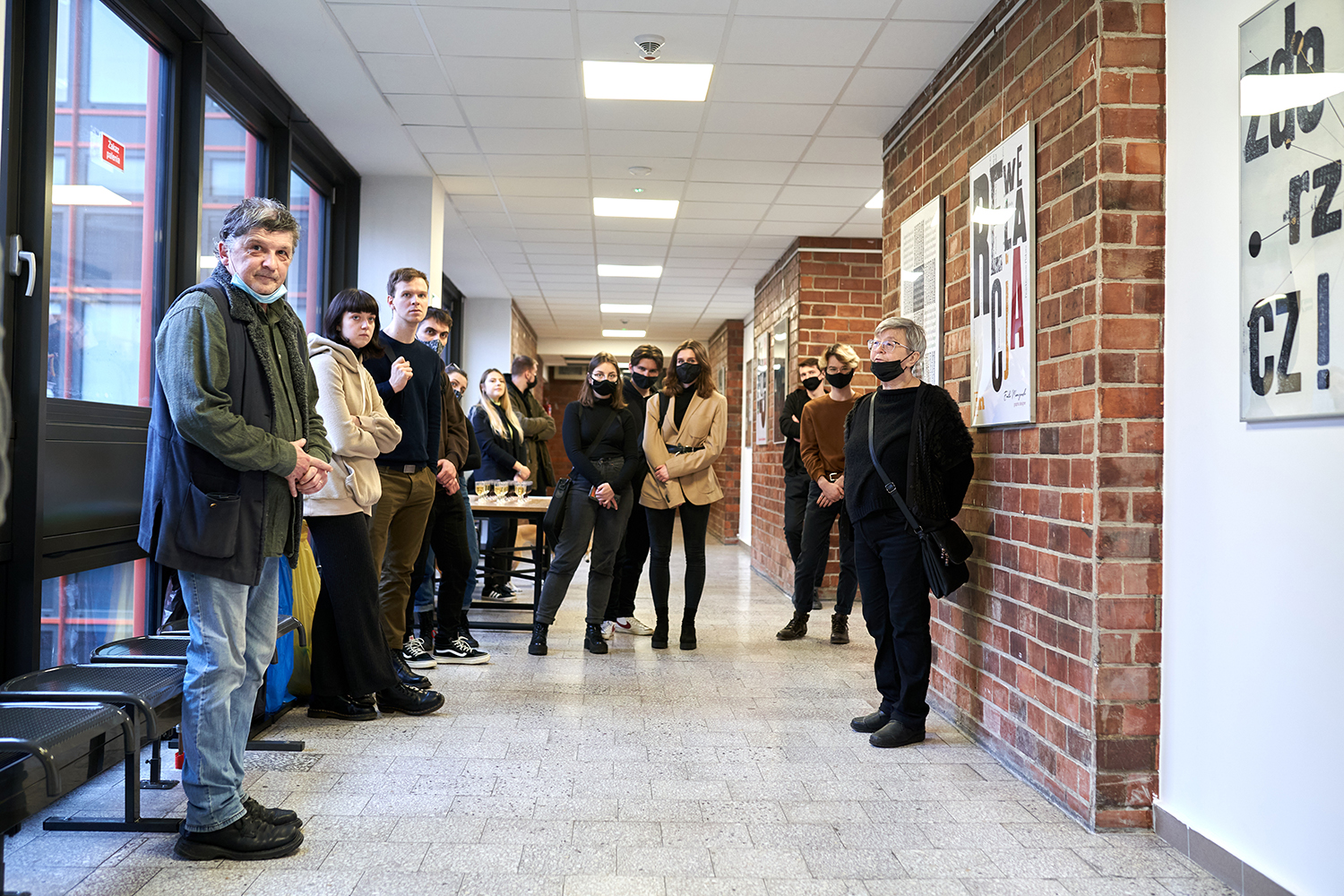 W galerii  101/121 ASP w Łodzi, na tle ceglanej ściany i wiszących tam plakatów, stoi kobieta w czarnym ubraniu, a wokół niej studenci i inni zainteresowani wystawą. Większość osób ma na twarzach ochronne maseczki.