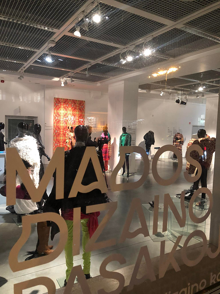 Przez szybę z beżowym napisem MADOS DIZAINO zaglądamy do wnętrza jasnej i przestronnej galerii. Są tam czarne postaci manekinów, które eksponują ubiory w wielu kolorach, oraz panele żakardowych i drukowanych tkanin.
