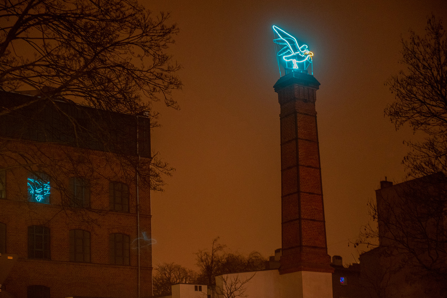 Ten sam neon, lecz pokazany w szerszym ujęciu, z pozycji widza stojącego znacznie niżej niż opisywany obiekt. Również widzimy go nocą, lecz tym razem, niewidoczne zza wysokich budynków  światła miasta, czynią niebo znacznie jaśniejszym. W powstałej łunie odczytujemy też wyraźne kształty zabudowań i komina, który wydaje się pełnić tu rolę postumentu czy  kolumny wznoszącej chwałę postaci uwiecznionej pomnikiem. Jest nią właśnie świetlista mewa.\nZdjęcie podpisano: Fot. Paweł Lacheta/ UMŁ