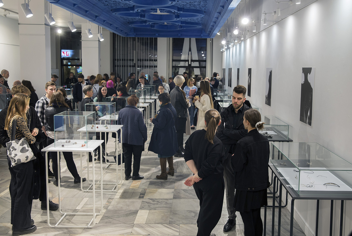 Ujęcie wykonane z głębi wnętrza galerii. Gabloty z biżuterią stoją po środku oraz pod ścianami,  w kilku długich rzędach. Tłumnie przybyli na wernisaż zwiedzający oglądają eksponaty.
