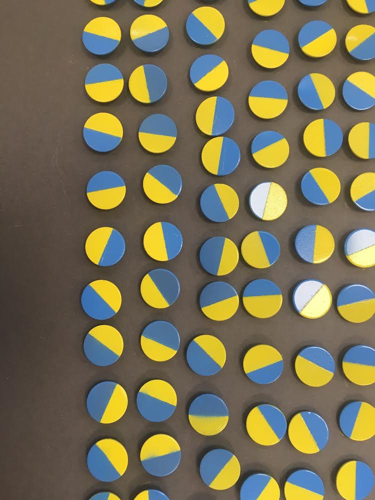 Wiele takich okrągłych tarcz upiętych gęsto obok siebie. Każda z nich w połowie jest błękitna, w połowie żółta. Na pokazanej w kadrze przestrzeni zgromadzono ich kilkadziesiąt. 