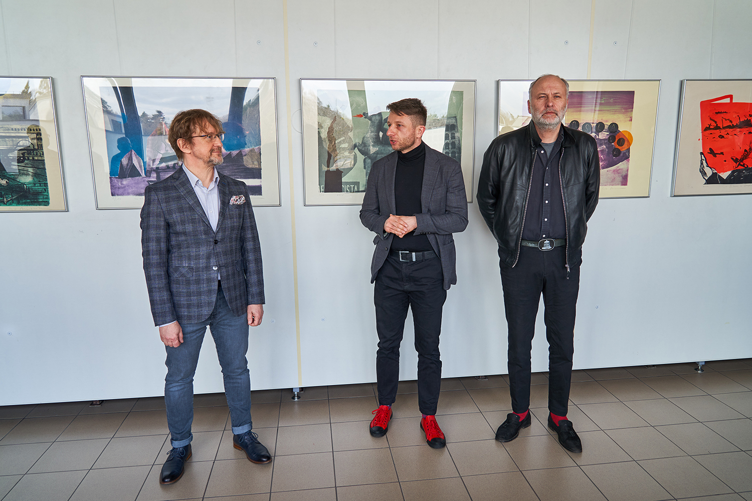 W galerii, na tle  kilku wiszących kolorowych prac, stoją trzej mężczyźni. Wśród nich jest Rektor uczelni, dr Przemysław Wachowski oraz autor wystawy.